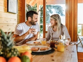 Dos adultos compartiendo desayuno en una mesa de madera dentro de una cabaña iluminada por luz natural, con jugo de naranja, tostadas y fruta fresca, en un ambiente cálido y acogedor.