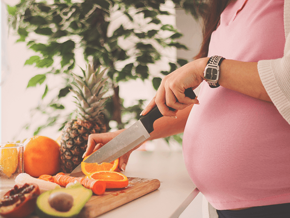 Pregnant woman slicing an orange by a kitchen counter full of fruit.