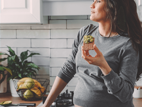 Dreamy looking pregnant woman, standin on her kitchen eating an avocado toast