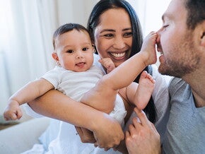 Mamá carga bebé sonriendo. Bebé tiene el pie en la nariz de papá.