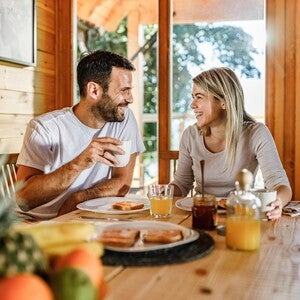 Dos adultos compartiendo desayuno en una mesa de madera dentro de una cabaña iluminada por luz natural, con jugo de naranja, tostadas y fruta fresca, en un ambiente cálido y acogedor.